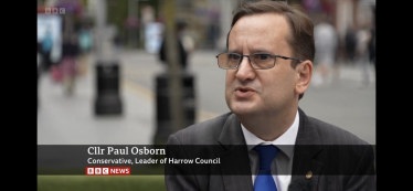 Paul Osborn in Harrow Town Centre wearing a suit, white shirt and a blue tie. In the background are people shopping.