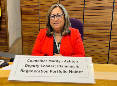 Cllr Marilyn Ashton sat in the old Council Chamber dressed in a red blazer looking at the camera and smiling. In front of her is her name plaque which reads 'Councillor Marilyn Ashton: Deputy Leader, Planning & Regeneration Portfolio Holder