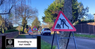 A photo showing a warning sign for road works. In the background are workmen working on a road. There are trees and houses either side of the road.